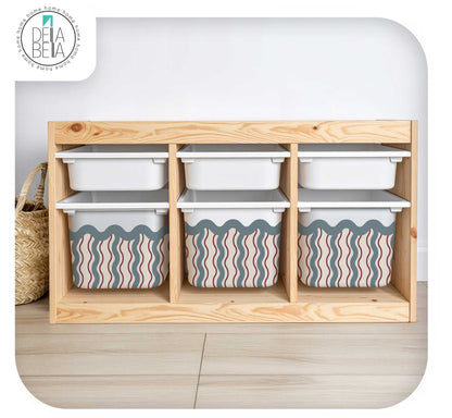 A light natural trofast storage organizer with six cubby shelves holding white plastic bins, with two lower bins featuring a pink, beige, and gray wavy stripe pattern. The organizer is set against a white wall and light wood floor, with a woven basket on the left.